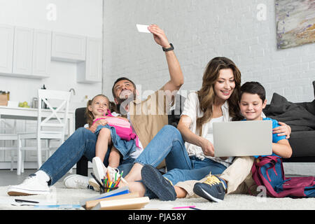 Lächelnd Vater und Tochter unter selfie auf Smartphone, während Mutter und Sohn mit Laptop zusammen zu Hause Stockfoto