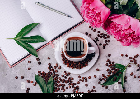 Schwarzen Kaffee in einer Tasse auf einem Tisch mit Pfingstrosen Notepad und Kaffeebohnen Draufsicht Stockfoto