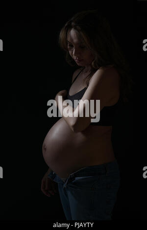 Eine schwangere asiatische Frau zur Seite stellen, nach unten schaut. Eine dunkle künstlerische Portrait. Stockfoto