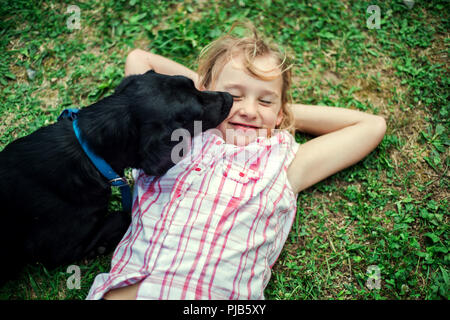 Ein kleines Mädchen liegen auf dem Rasen, ein schwarzer Hund leckt ihr Gesicht. Stockfoto