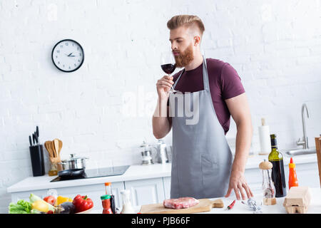 Gut aussehender bärtiger junger Mann in Schürze Weinprobe Rotwein beim Kochen in der Küche Stockfoto