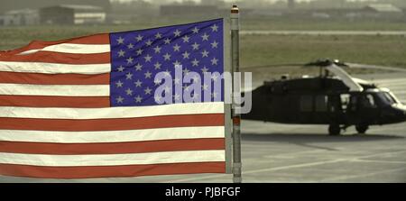 (Bagram, Afghanistan) die amerikanische Flagge im Wind Wellen mit einer UH-60 Blackhawk Hubschrauber zu Task Force Schatten auf dem Flug zugewiesene Leitung geparkt. Juli 5, 2018. Stockfoto