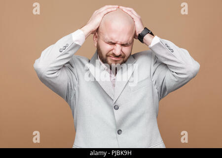 Kopfschmerzen und Schmerzen. Portrait von mittleren Alters kahlen bärtigen Geschäftsmann in Hellgrau Anzug stehen und seine schmerzhaften Kopf. indoor Studio shot, isol Stockfoto