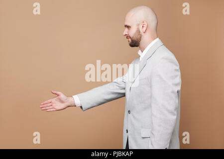 Die Hand schütteln und Gruß. Profil Seitenansicht Portrait von mittleren Alters kahlen bärtigen Geschäftsmann in Hellgrau Anzug zu stehen und die Hand. indoor Studio Stockfoto