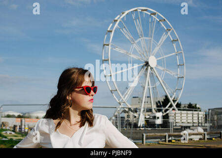 Ein Mädchen stand vor einem Riesenrad mit roter Sonnenbrille für ein Modeschießen. Stockfoto