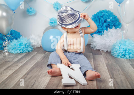 Portrait von cute adorable kaukasische Baby Junge mit blauen Augen barfuß in Hose mit Hosenträgern, unter Hut fallen versteckt, sitzen auf den Holzboden in s Stockfoto