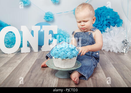 Portrait von cute adorable Blonde kaukasische Baby Junge mit blauen Augen in Jeans Overall, seinen ersten Geburtstag mit Gourmet Kuchen Feiern, Briefe und Stockfoto