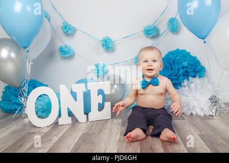 Portrait von adorable kaukasische Baby boy in dunklen Hosen und blaue Fliege seinen ersten Geburtstag mit Buchstaben ein und Luftballons feiern, sitzen auf den Woode Stockfoto