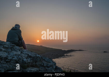 St. Ives, England - Mai 2018: Mann sitzt auf einem Felsen und bewundern Sie den Sonnenuntergang über der Cornwall, Cornwall Stockfoto