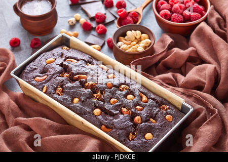 Close-up von Schokolade Buchweizen Pound Cake mit Muttern und Himbeeren in eine Backform auf einer konkreten Tabelle mit Tasse Kaffee und braun Gewebe, v Stockfoto