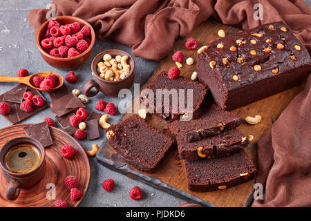 Close-up von Schokolade Buchweizen Pound Cake mit Muttern und Himbeeren in Scheiben schneiden auf einem Holzbrett auf einer konkreten Tabelle mit Tasse Kaffee und Braun Stockfoto