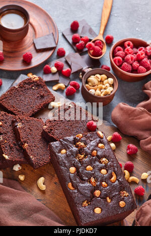 Schokolade Buchweizen Pound Cake mit Muttern und Himbeeren in Scheiben schneiden auf einem Holzbrett auf einer konkreten Tabelle mit Tasse Kaffee und braunen Tuch, Verti Stockfoto
