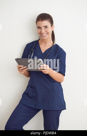 Junge Frau Arzt oder Krankenschwester in Blau scrubs Holding ein Tablet PC vor einer weißen Wand Stockfoto