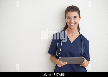 Junge Frau Arzt oder Krankenschwester in Blau scrubs Holding ein Tablet PC vor einer weißen Wand Stockfoto