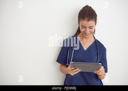 Junge Frau Arzt oder Krankenschwester in Blau scrubs Holding ein Tablet PC vor einer weißen Wand Stockfoto