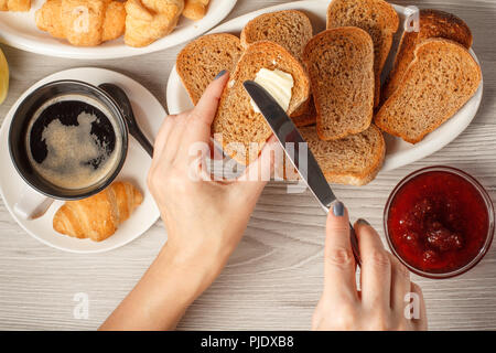 Weibliche Hände Verbreitung Butter auf Toast mit Tasse schwarzen Kaffee und Croissant, Brot, Glasschale mit Marmelade auf dem Hintergrund. Ansicht von oben. Essen und bevera Stockfoto