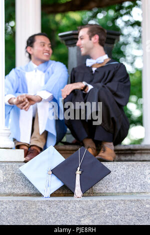 Zwei männliche Hochschulabsolventen sitzen auf der Treppe im Gespräch mit mortarboards im Vordergrund positioniert Stockfoto