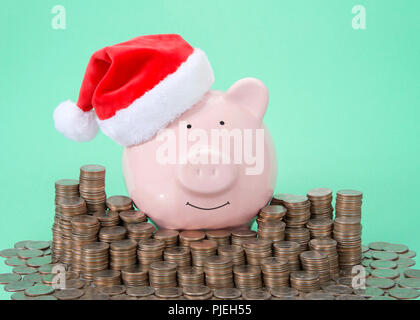 Rosa Sparschwein Tragen eines Santa Hut lächelnd mit Blick auf Viewer durch Stapel von Münzen, grüner Hintergrund umgeben. Stockfoto