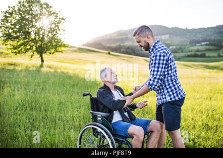 Ein erwachsener hipster Sohn mit dem Vater im Rollstuhl auf einen Spaziergang in der Natur bei Sonnenuntergang. Stockfoto