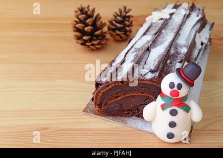 Schokolade Yule Log Kuchen für Weihnachten oder Buche de Noel mit einem niedlichen Schneemann Marzipan und zwei trockene Pin Kegel auf Hellbraunem Holz- Tabelle Stockfoto
