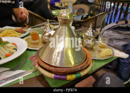 Traditionelle marokkanische Gericht namens Tagine in lokaler Sprache auf Tisch im Restaurant in Marrakesch, Platten mit anderen Mahlzeiten, die Hand der Person, Gläser mit Tee. Stockfoto