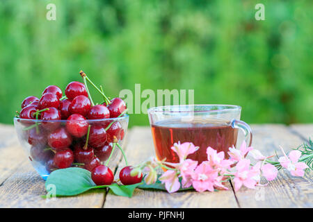 Rote reife Kirsche Früchte mit dem Stiel in Glasschale und Tasse Tee auf alten hölzernen Brettern. Selektiver Fokus auf Kirschen Stockfoto