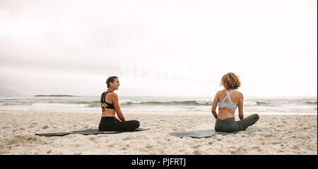 Rückansicht zweier Frauen in Fitnesskleidung, die Yoga üben, am Strand sitzen. Fitness-Frauen sitzen auf Yogamatten und machen Yoga mit Blick auf das Meer. Stockfoto