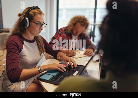 Computer programmierer bei Coworking Space in technischer Startupservice für die Arbeit im Büro. Frau mit Kopfhörern Codierung auf Laptop mit Kollegen rund um den t Stockfoto