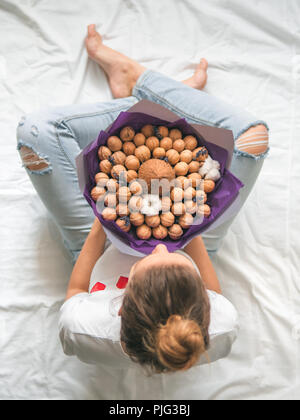 Junge Frau sitzt auf dem Bett mit Kokos und Walnüsse essen Blumenstrauß in den Händen. Blick von oben auf die Jungen hipster Frau in zerrissenen Jeans am Knie mit ungewöhnlichen Stockfoto