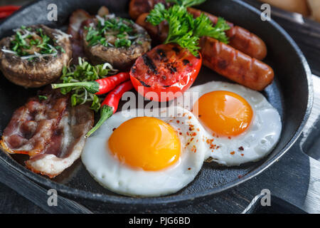 Nahaufnahme des Englischen Frühstück in einer Pfanne mit Spiegelei, Würstchen, Speck, Pilzen, Marmelade und Orangensaft auf dunklem Stein Hintergrund Stockfoto