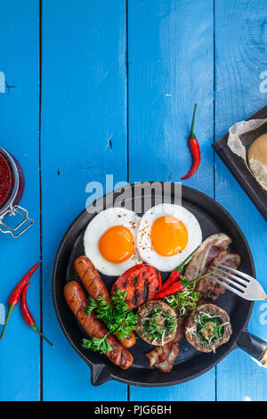 Englisches Frühstück in einer Pfanne mit Spiegelei, Würstchen, Speck, Pilzen, Marmelade und Orangensaft auf hölzernen Background Copy space Stockfoto