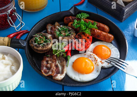 Ansicht schließen Englisches Frühstück in einer Pfanne mit Spiegelei, Würstchen, Speck, Pilzen, Marmelade und Orangensaft auf hölzernen Hintergrund Stockfoto