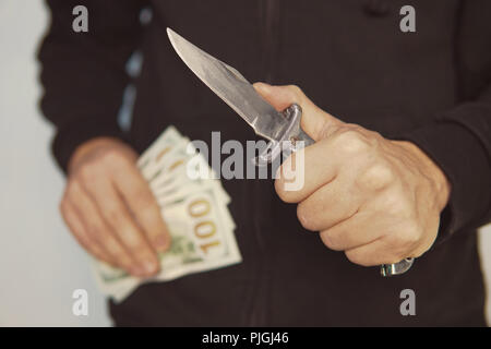 Räuber mit Messer in der Hand und Amerikanisches Geld. Bandit zieht seine Pocket Taschenmesser auf Bedrohungen. ein Räuber mit einem Messer in der Kapuze. Das Konzept o Stockfoto