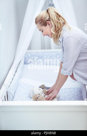 Gerne schwangere Frau bereitet ein Kinderzimmer für Ihr Baby, dekorieren eine Krippe mit Soft toys, junge weibliche genießen Schwangerschaft Zeit Stockfoto