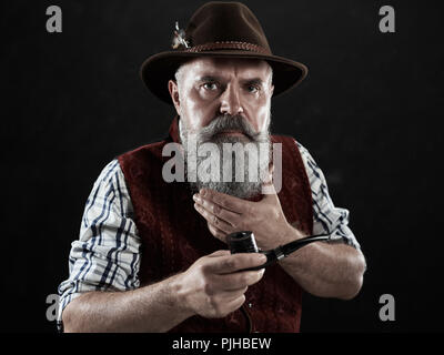 Dramatische Portrait von grauen Bärtigen älteren Mann in hat Tabak Pfeife. Blick auf die österreichischen und Tiroler, Bayerische alter Mann in nationaler Tracht im Retro-Stil. Stockfoto