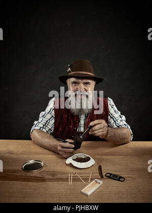 Die älteren bärtigen Mann am Tisch sitzen und das Verstopfen der Tabak in Rohr. Die männlichen Händen hautnah. Bayern. ein Mann in traditionelle bayerische oder Österreichische nationale Tracht gekleidet Stockfoto