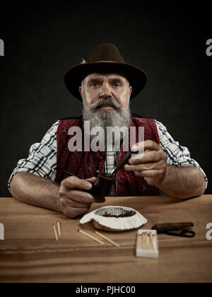 Die älteren bärtigen Mann am Tisch sitzen und das Verstopfen der Tabak in Rohr. Die männlichen Händen hautnah. Bayern. ein Mann in traditionelle bayerische oder Österreichische nationale Tracht gekleidet Stockfoto
