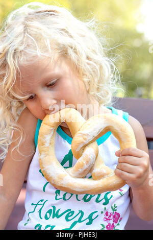 Adorable kleinen Mädchen auf der Bank sitzen mit Ihrem pretsel im Park. Blonde kleine Mädchen Essen im Freien. Stockfoto