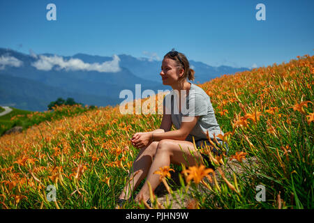 Junge Frau sitzt in den Bereichen orangen Blüten mit Bergen im Hintergrund Stockfoto