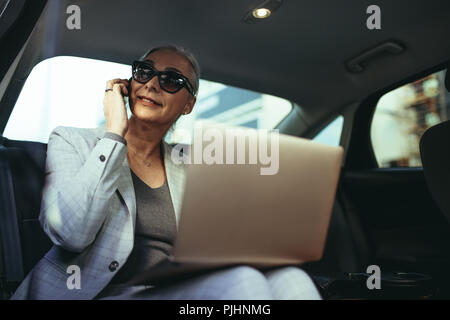 Weibliche Unternehmer mit Sitz in einem luxuriösen Auto saß auf dem Rücksitz mit Laptop und Suchen außerhalb des Fensters während Sie am Telefon sprechen. Ma Stockfoto