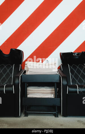 Zwei schwarze Ledersitze mit Waschbecken in einem barbershop Interieur mit Handtücher und Shampoo auf der Seite. Rot-weißen Streifen an der Wand. Stockfoto