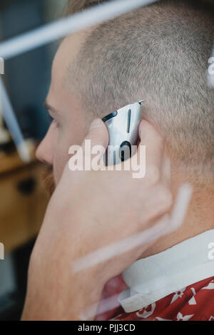 Ein junger gut aussehender bärtiger Mann in einem Friseursalon. Der Friseur Bürsten sein Haar und schneidet sie mit einem elektrischen Trimmer. Stockfoto