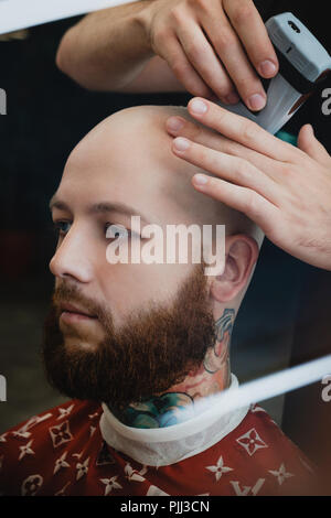 Ein gut aussehender Bärtiger skinhead Mann in einem Friseursalon. Der barbier rasiert seinen Kopf mit einem elektrischen Trimmer. Stockfoto