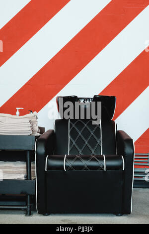 Zwei schwarze Ledersitze mit Waschbecken in einem barbershop Interieur mit Handtücher und Shampoo auf der Seite. Rot-weißen Streifen an der Wand. Stockfoto