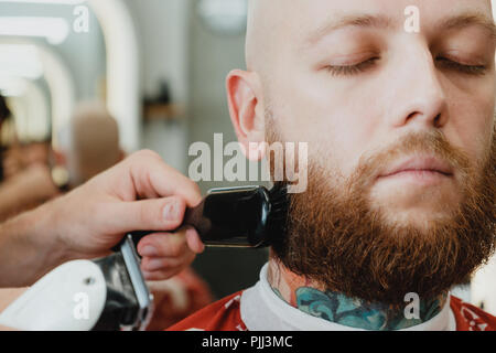 Ein gut aussehender Bärtiger skinhead Mann in einem Friseursalon. Die Friseur- schläge von der abgeschnittenen Haare mit einer Bürste. Stockfoto