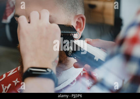 Ein junger gut aussehender bärtiger Mann in einem Friseursalon. Der Friseur Bürsten sein Haar und schneidet sie mit einem elektrischen Trimmer. Stockfoto