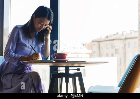 Asiatische Frau Gespräch am Handy. Junge Frau im Cafe sitzen und schreiben auf Notepad. Stockfoto