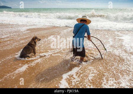 Ein Hund sitzt im Meer mit einem Fischer, während ein Fischer fischen Sand crab am Mai Khao Beach in der Nähe von Phuket Airport Stockfoto