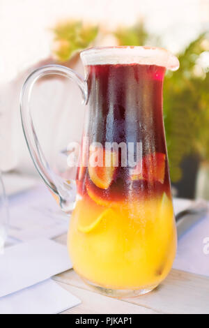 Hohes Glas kanne Krug mit Eis kalt gelbe und rote Sangria mit Kondensation auf dem Glas und Fruchtstücken innen auf einem Holz Tisch gesehen. Die Kanne hat eine Stockfoto