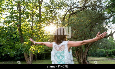 Junge Frau feiert die Morgen Sonne und Frühling Jahreszeit, während Sie im Freien steht im Garten bei Sonnenaufgang mit ausgebreiteten Armen, Ansicht von hinten. Stockfoto